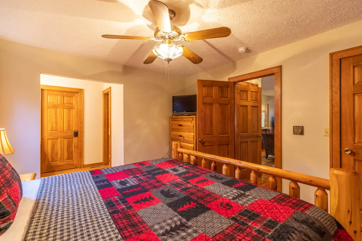 A comfortable bedroom showcasing plaid bedding, wooden furniture, and a ceiling fan. Sunlight filters through the window, enhancing the inviting atmosphere. The space is designed for peaceful relaxation and restful sleep.
