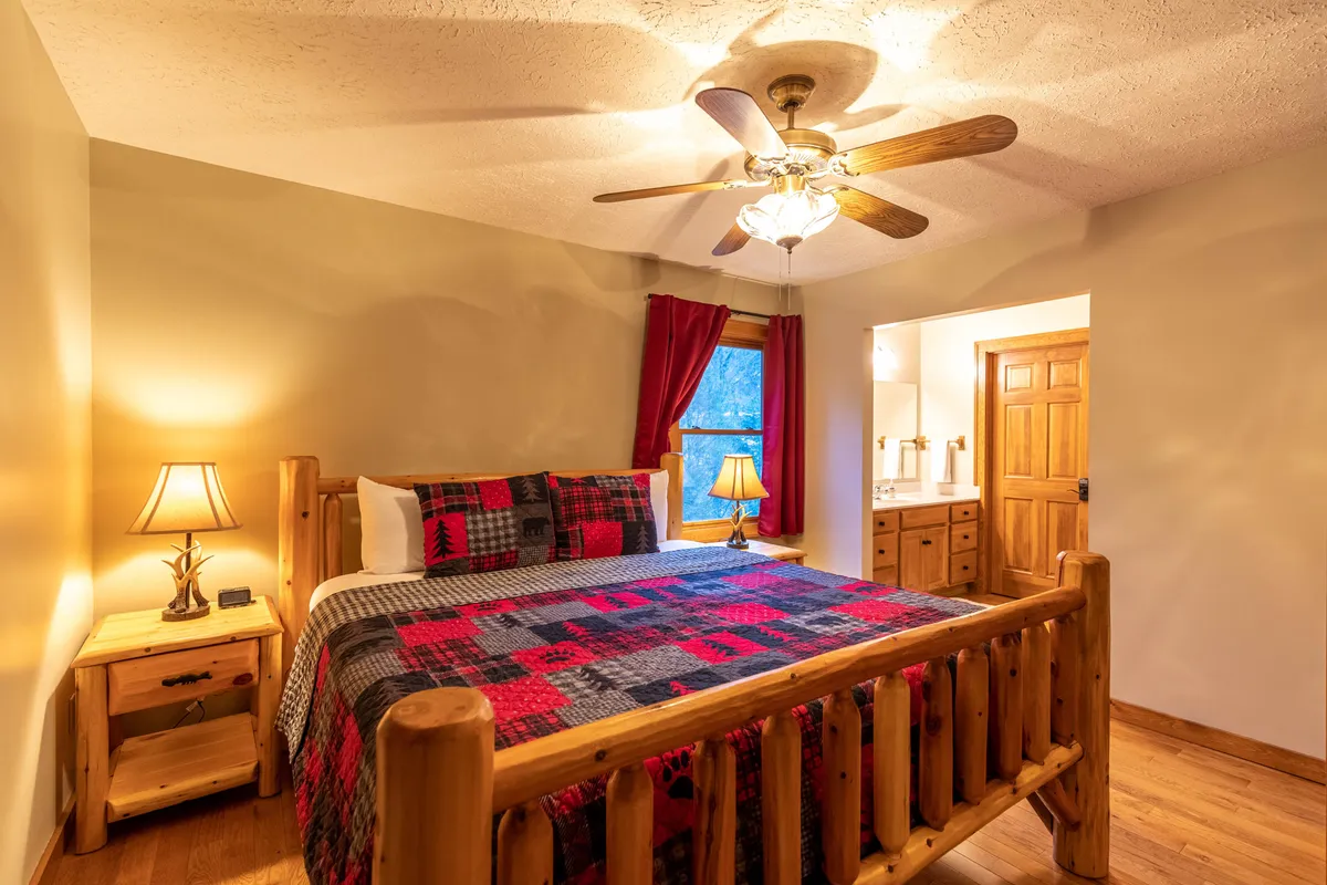 A warm, inviting bedroom featuring a rustic log bed with a plaid blanket, soft table lamps, and a window with curtains that lets in gentle natural light. The cozy details create an intimate, restful atmosphere ideal for relaxing or reading before bed.