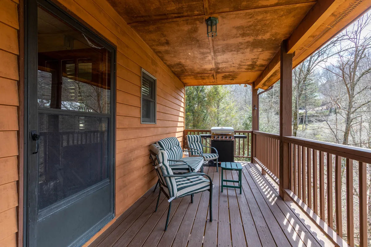 The image captures a casual outdoor living space with striped chairs, a table set for dining, a grill, and a wooden railing overlooking the surrounding landscape. The porch setting invites guests to enjoy meals, relax, and socialize outdoors, emphasizing the connection to the natural mountain environment.