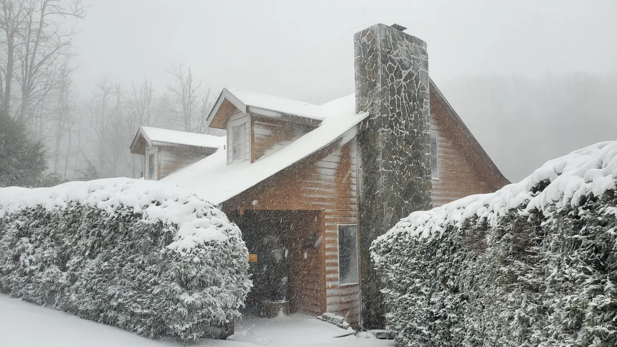 A charming exterior shot of the house in winter, with a snow-covered roof, stone chimney, and evergreen hedges. The snow creates a tranquil mountain setting, reinforcing the need for seasonal readiness.