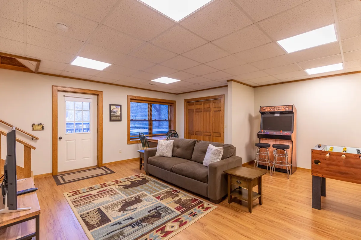 This image features a casual and inviting game room with a comfortable sofa, a vibrant arcade game, foosball table, large rug, and windows letting in natural light. The space is clearly set up for fun and relaxation, with room for socializing, playing games, and unwinding.
