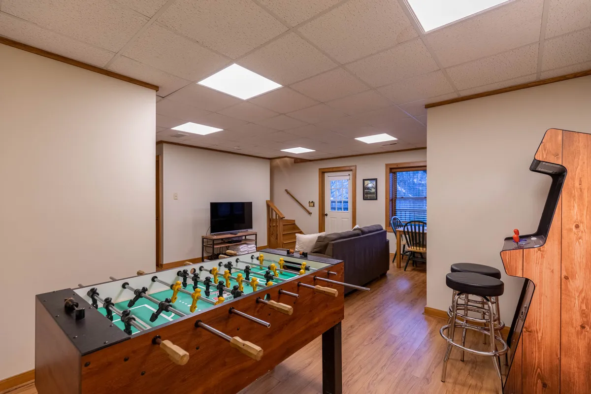 A playful and relaxed entertainment space with a foosball table as the focal point, comfortable seating, a television, and an arcade machine. The setup encourages competition, relaxation, and group activities, with all the key entertainment elements visible.
