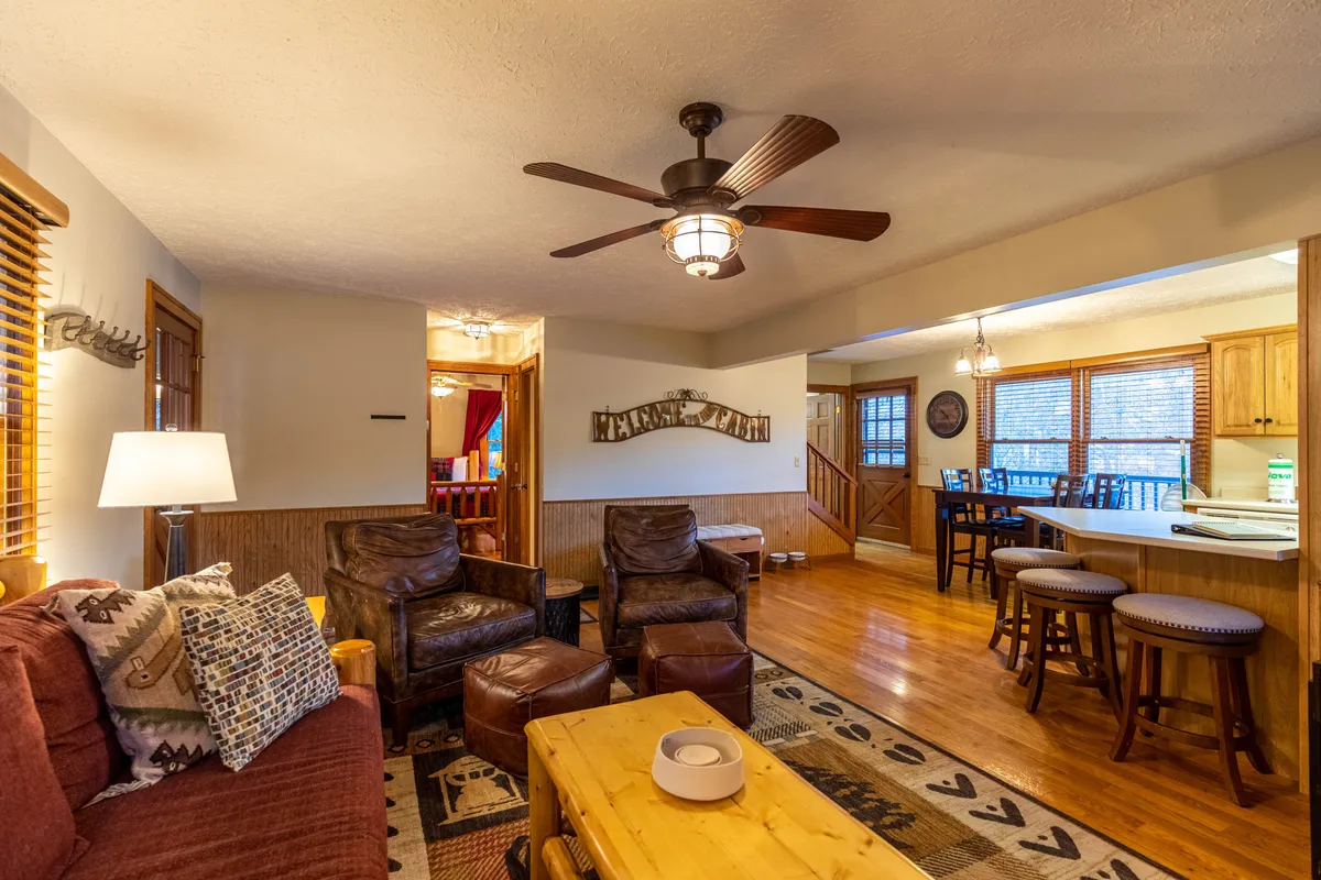 This image displays a comfortable seating area with rustic decor and a wooden coffee table. The living space opens up toward the kitchen bar area, giving a sense of openness and warmth. The large windows and inviting setup suggest a cozy interior that feels connected to the outdoors.