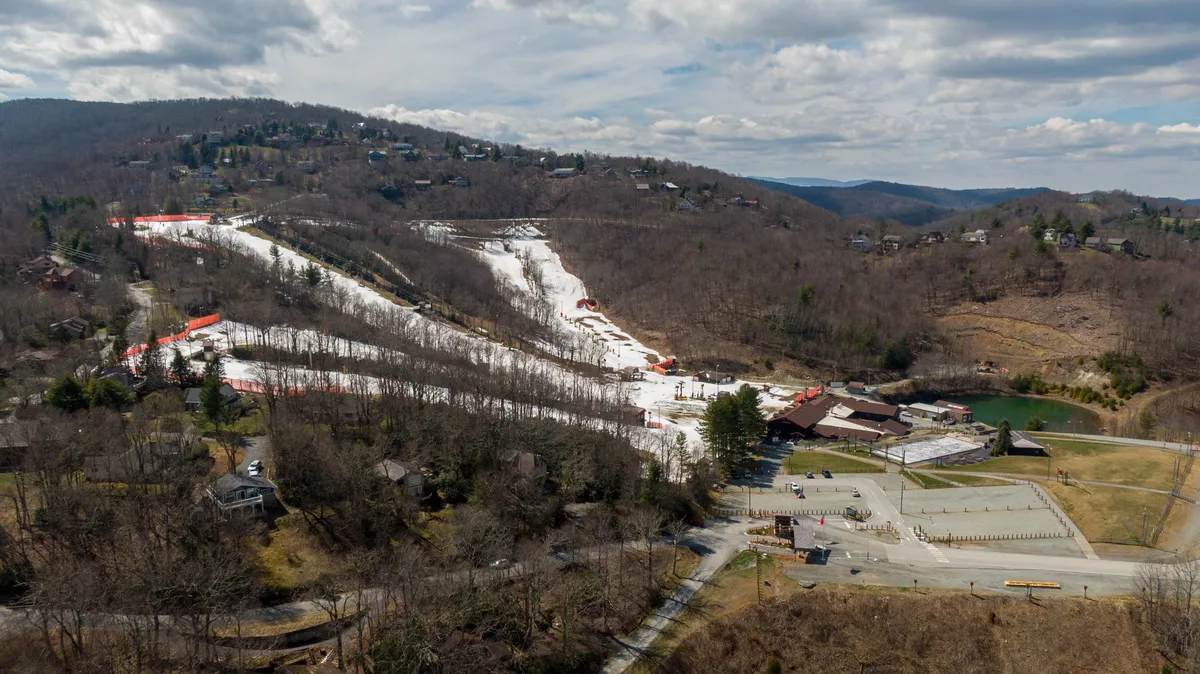 A sweeping view of the Appalachian Ski Mountain area, featuring ski slopes, the lodge, nearby homes, and a parking area. Snow dusts the slopes and residential rooftops, creating a sense of adventure and seasonal excitement.
