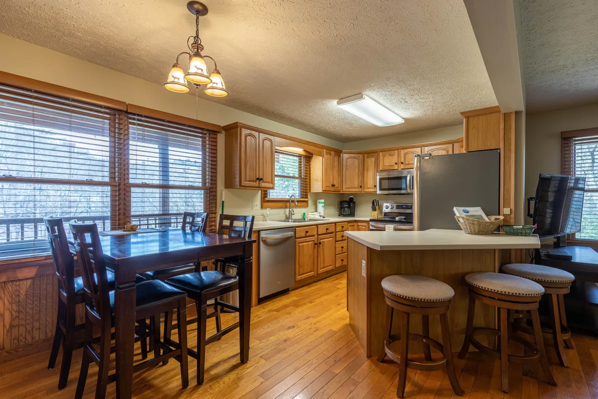 This photo highlights the wooden dining table and kitchen peninsula with stools, surrounded by cabinetry and natural light from nearby windows. It conveys a space designed for shared meals, breakfasts, and social gatherings, with a clear view into the kitchen, reinforcing the sense of openness and flow.