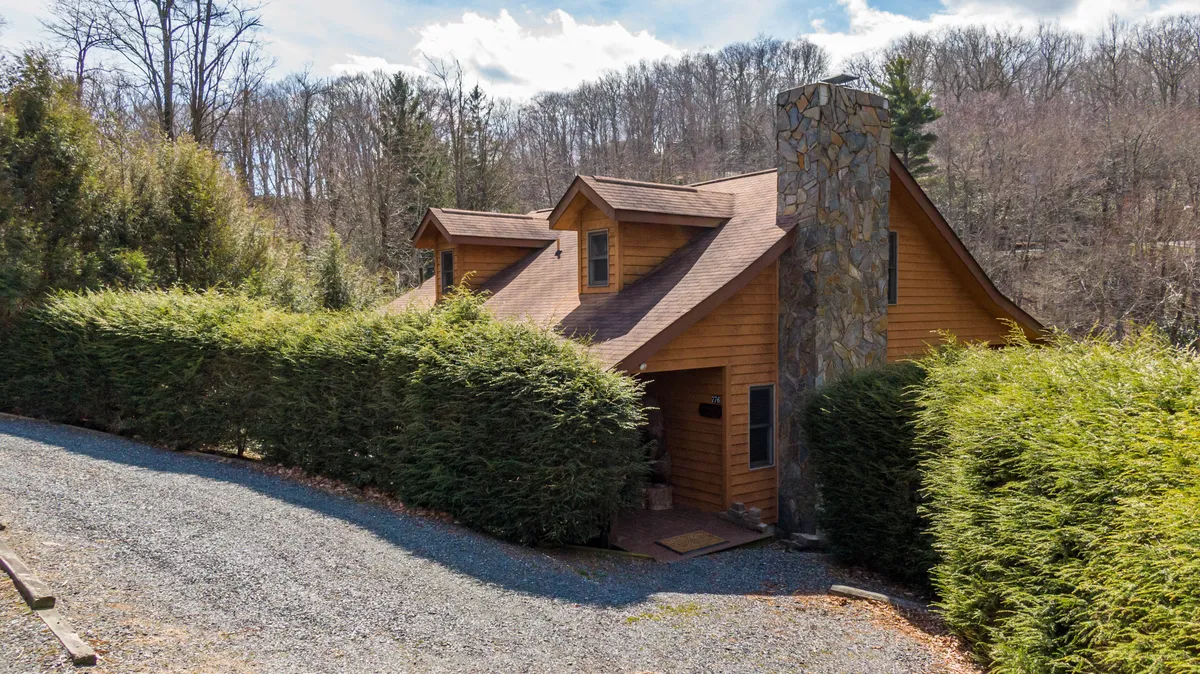 The rustic wooden cabin with a stone chimney nestles in lush greenery, surrounded by trees. The inviting front porch offers a cozy spot to relax and connect with nature, while the setting hints at the proximity to trails and outdoor spaces ideal for exploration with family and pets.